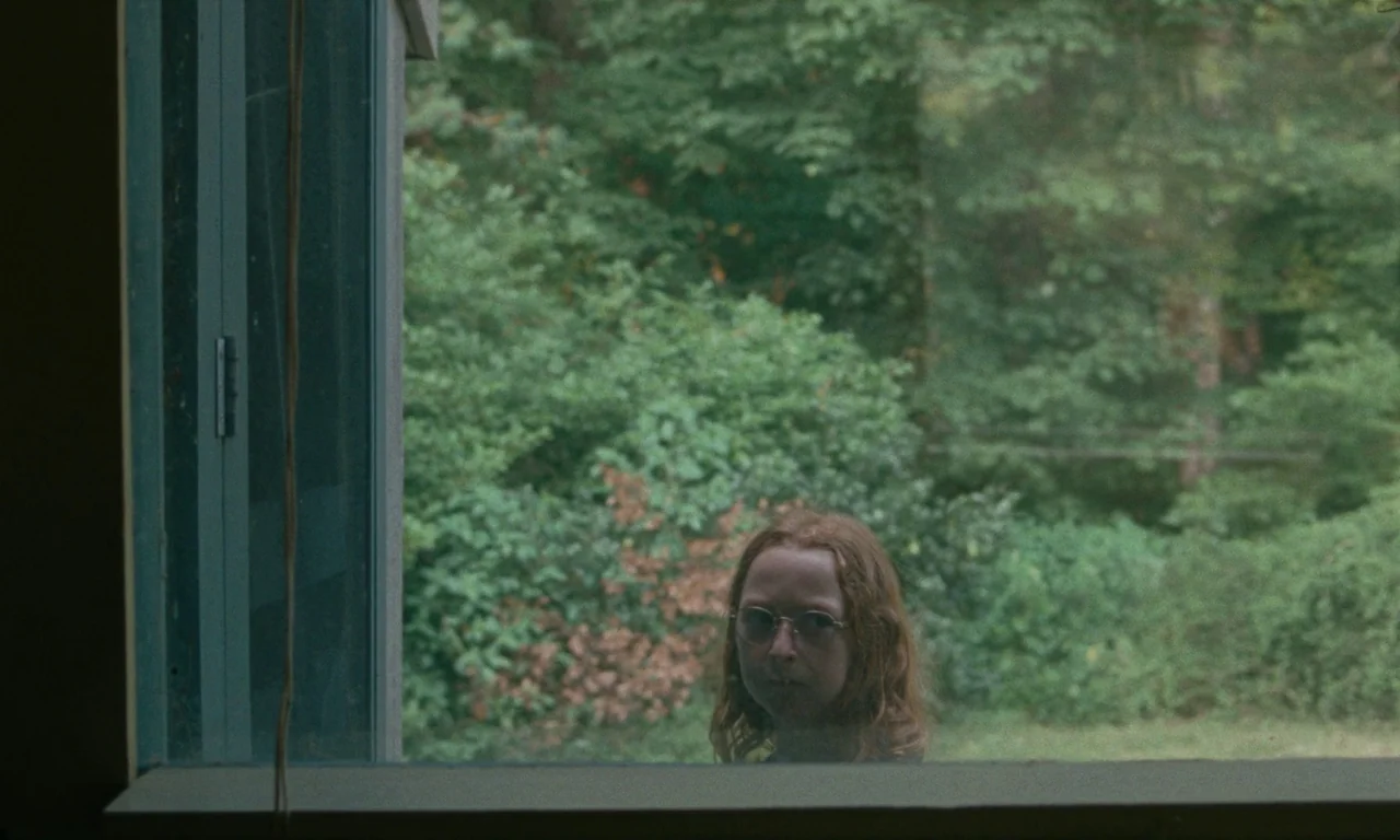 Janet Planet (2023) movie still: medium — A person stands outside looking through a window into an interior space. The backdrop is a densely wooded…