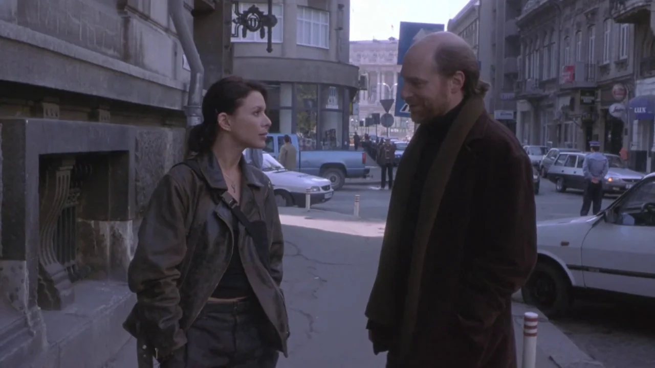Hellraiser: Deader (2005) movie still: medium — A city street corner with classic European-style architecture and parked automobiles nearby. A woman…