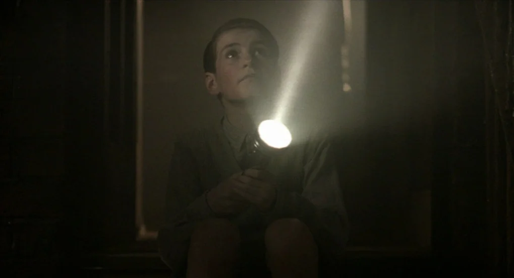 The Long Day Closes (1992) movie still: medium — A dark, dimly lit indoor space with blurred architectural shapes in the background. A young boy…