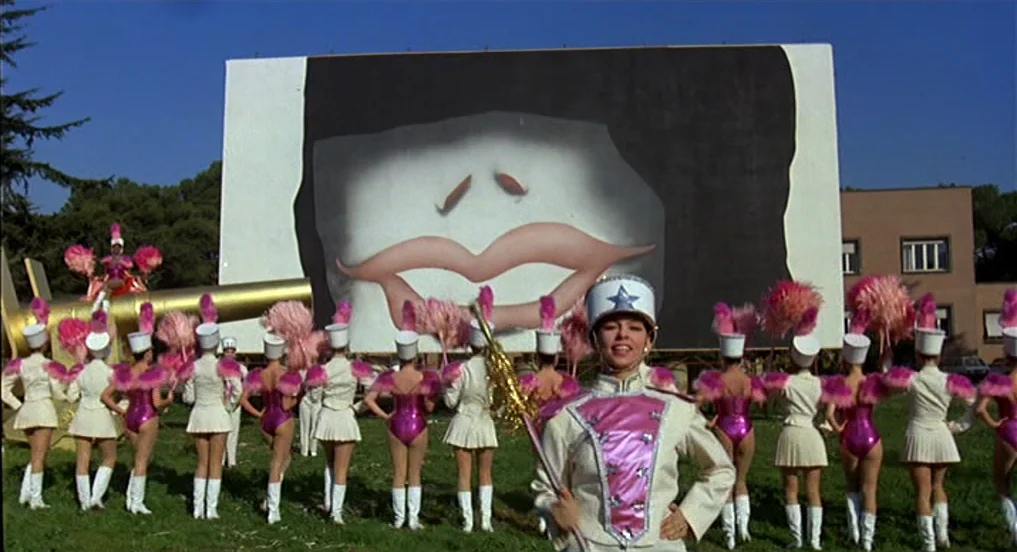 Intervista (1987) movie still: wide — A group of women in marching uniforms stand in a grassy field before a screen. A large cinematic screen…