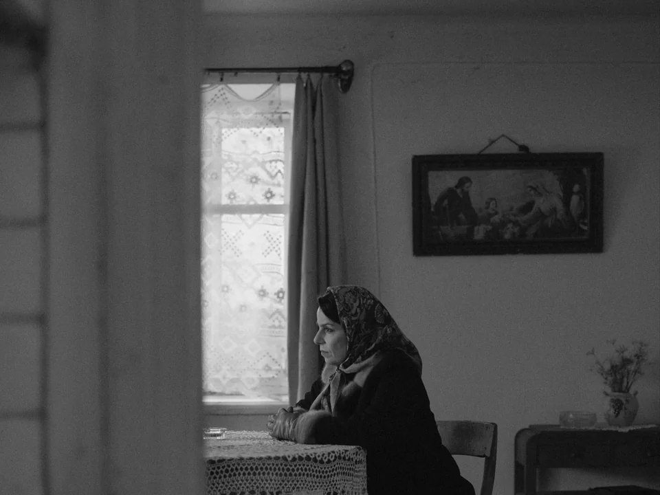 Ida (2013) movie still: medium — A plain interior with white walls features a lace-covered table and a religious painting. A woman in a dark…