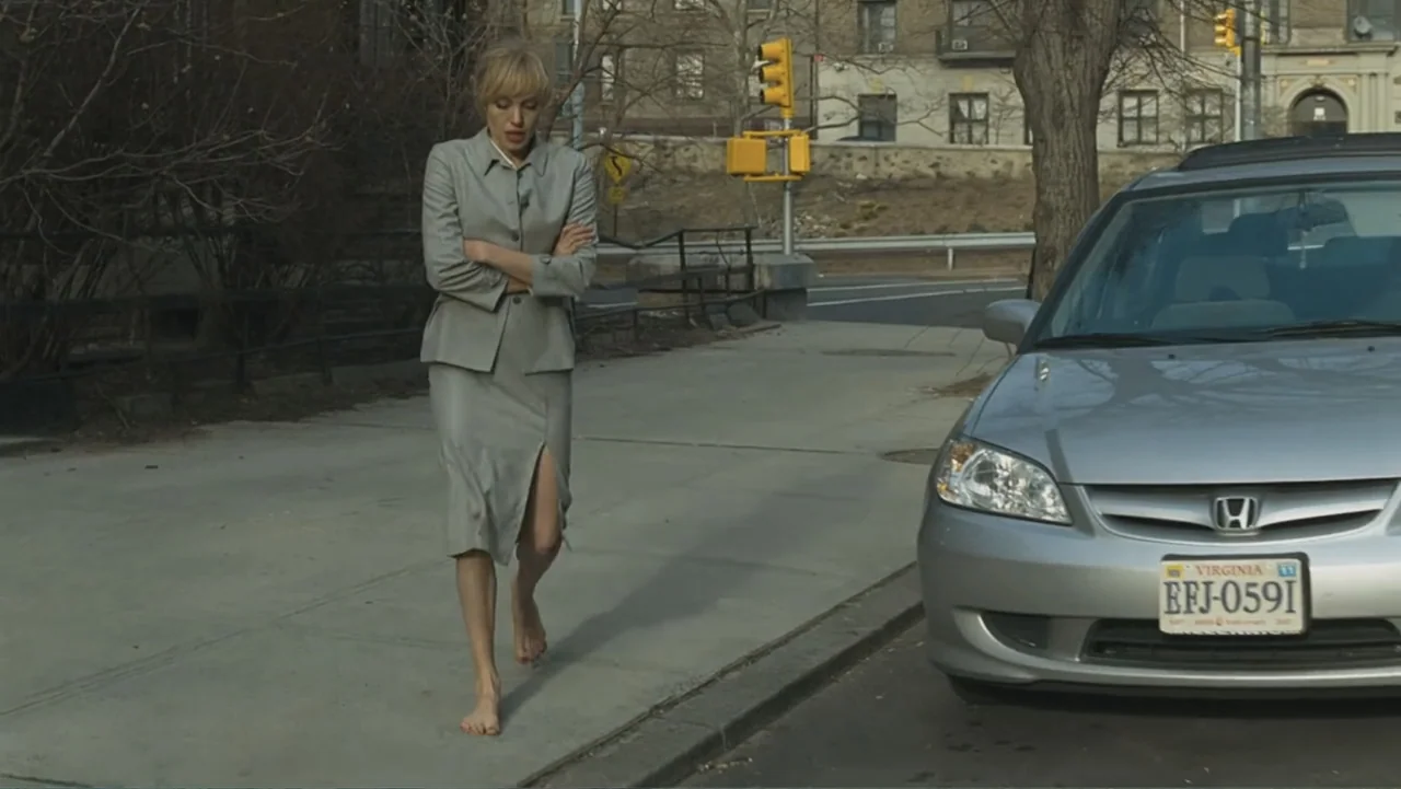 Salt (2010) movie still: fullbody — A sidewalk borders a road with a parked silver car and urban background. A woman in a gray skirt suit walks…