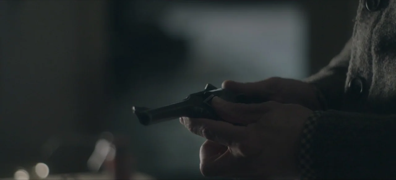 We Go In at Dawn (2020) movie still: closeup — A dark indoor space contains a person holding a small vintage firearm. The person's hands are visible…