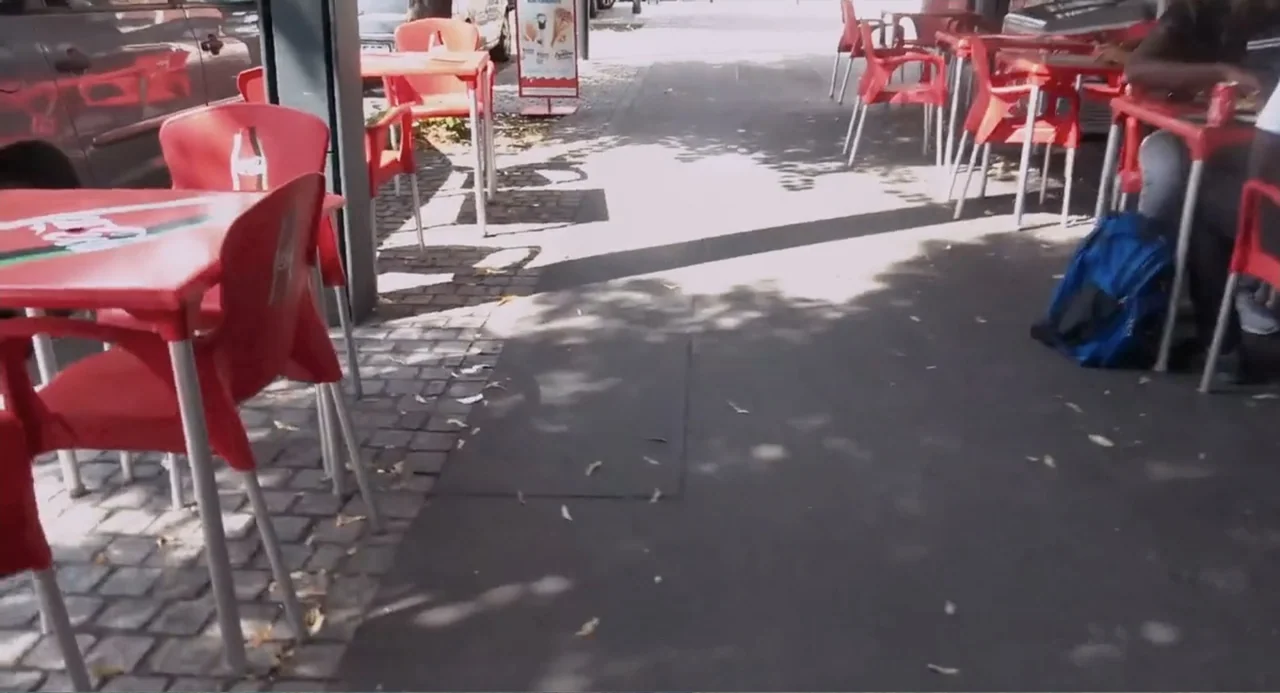 A Dramatic Film (2019) movie still: wide — A sidewalk cafe features bright red plastic chairs and matching tables arranged on cobblestone. A blue…