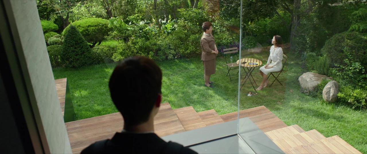 Parasite (2019) movie still: wide — A lush backyard features a wooden terrace and a small bistro set on grass. Two women stand and sit by a table…