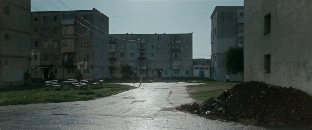Graduation (2016) movie still: establishing — A desolate residential courtyard features multi-story Soviet-style apartment buildings with peeling…