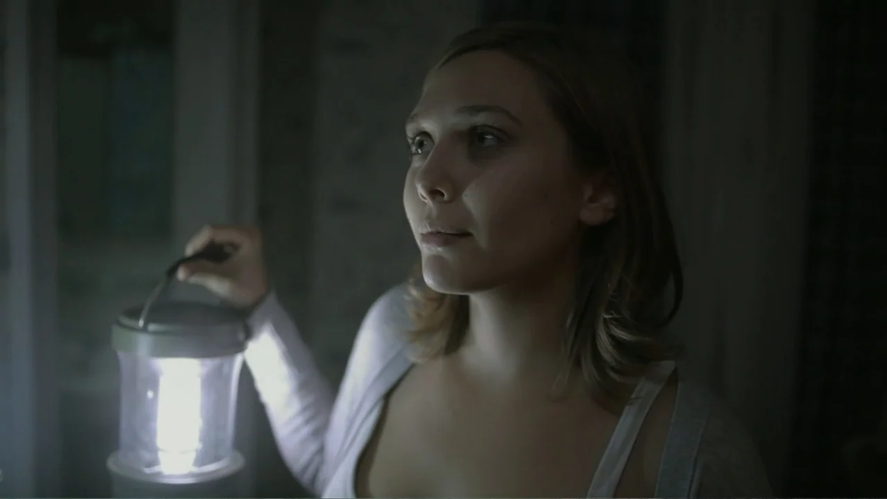 Silent House (2011) movie still: medium — A dimly lit interior space with the subject standing in darkness. A young woman holds a bright electric…