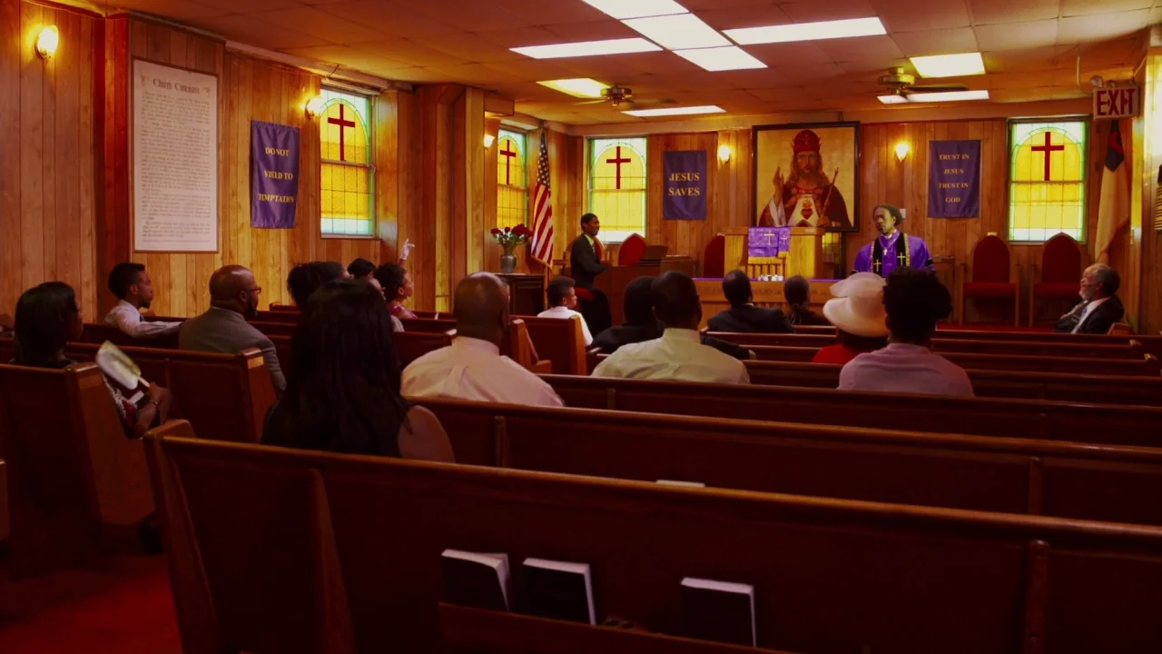 Red Hook Summer (2012) movie still: wide — A wood-paneled community church features rows of pews facing a small altar area. A pastor stands at the…