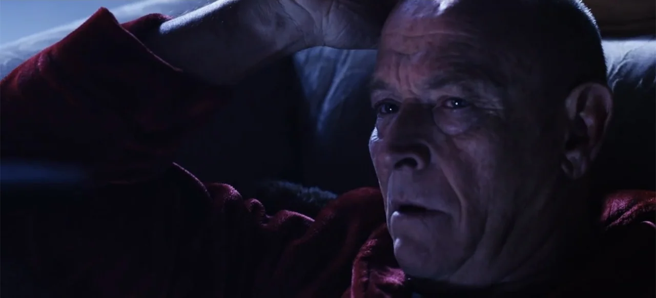 The Russian Bride (2019) movie still: closeup — An older man is positioned in a dimly lit indoor space at night. He is wearing a dark red robe and…