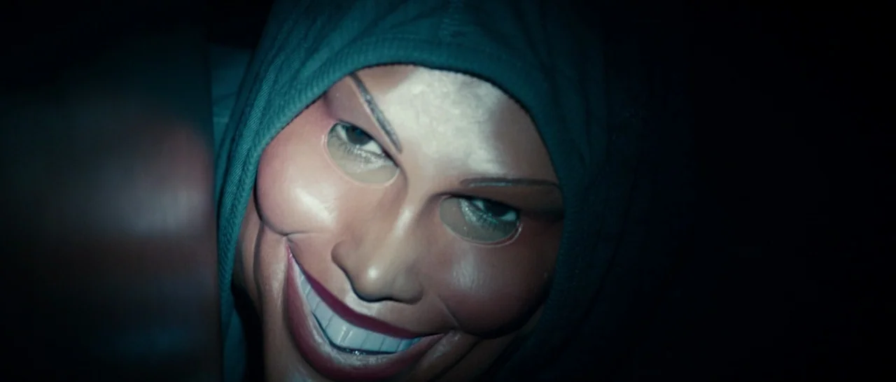 The Purge (2013) movie still: closeup — A darkened interior space features a person wearing a plastic mask peeking through a gap. The subject stares…