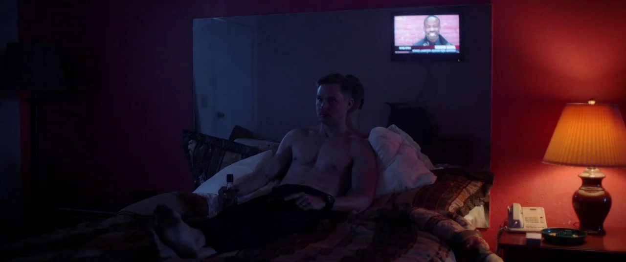 Blindfire (2020) movie still: medium — A dimly lit motel room features red walls and a large mirror above the bed. A shirtless man sits on the bed…