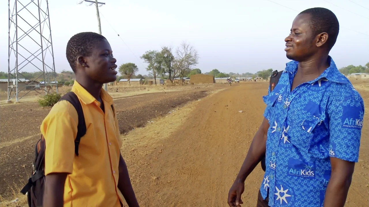 The Money Stone (2018) movie still: medium — A dusty dirt road in a rural landscape features a tall metal transmission tower. Two men wearing…