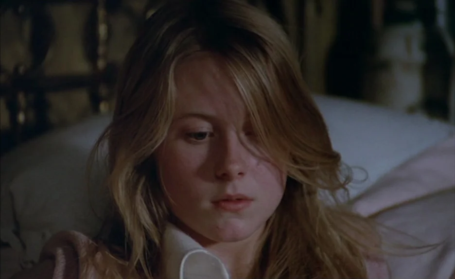 Black Moon (1975) movie still: closeup — A dimly lit indoor space features a young woman resting on a bed. The subject lies against pillows with her…