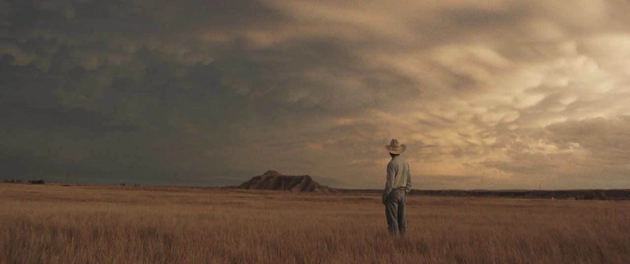 The Rider (2017) movie still: wide — A vast, open grassy plain stretches toward a solitary, eroded mesa under dramatic clouds. A man wearing a…