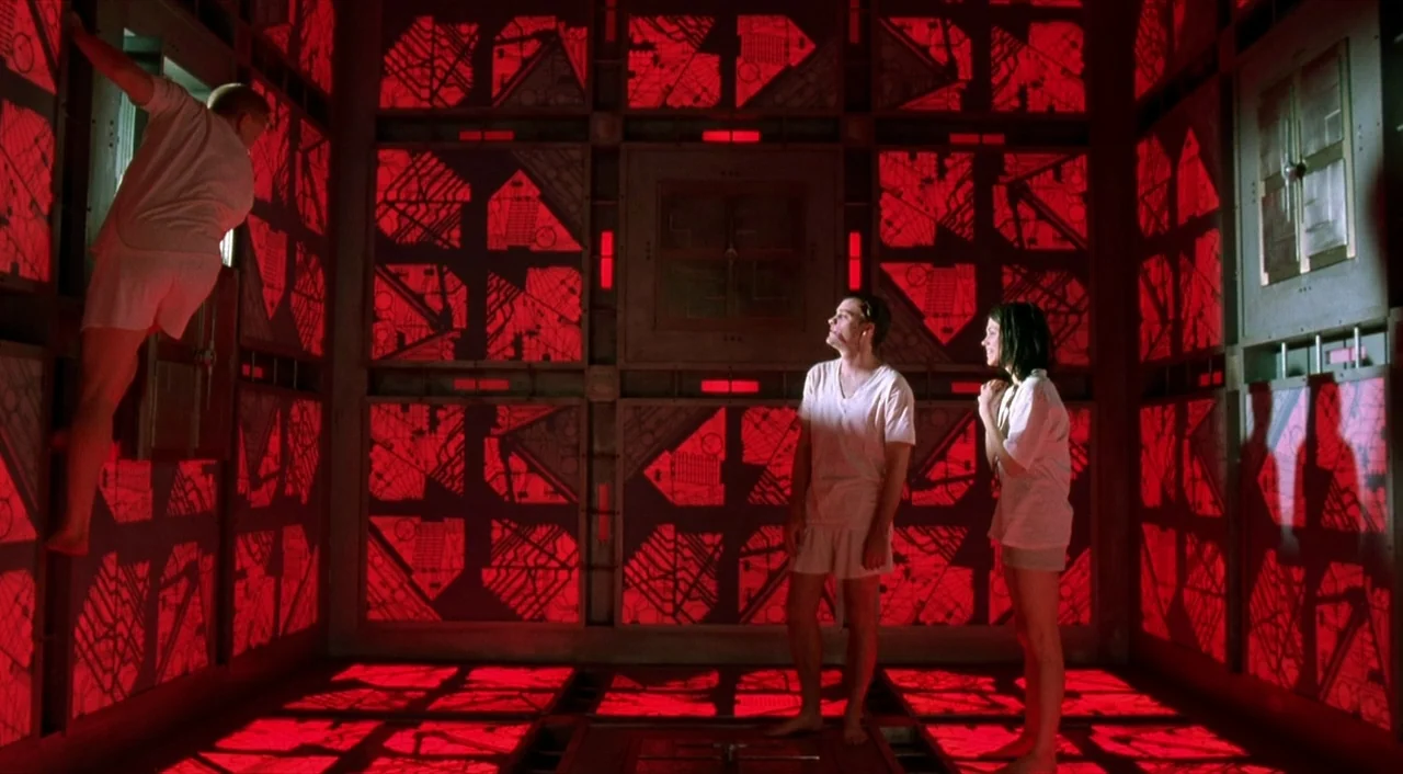 Cube (1997) movie still: wide — A confined metallic space features intricate geometric red patterns glowing on every surface. Three individuals are…