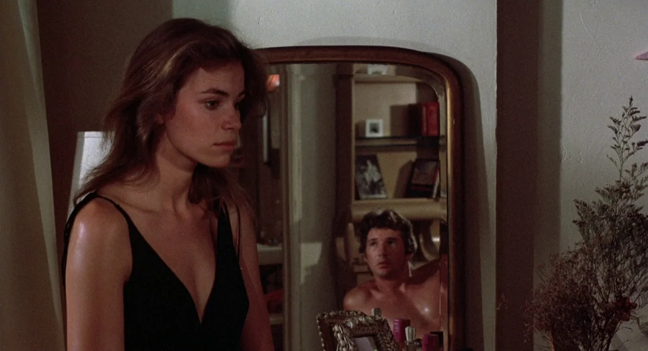 Breathless (1983) movie still: medium — A woman stands in a dimly lit room near a vanity mirror. Her partner is visible as a reflection in the…
