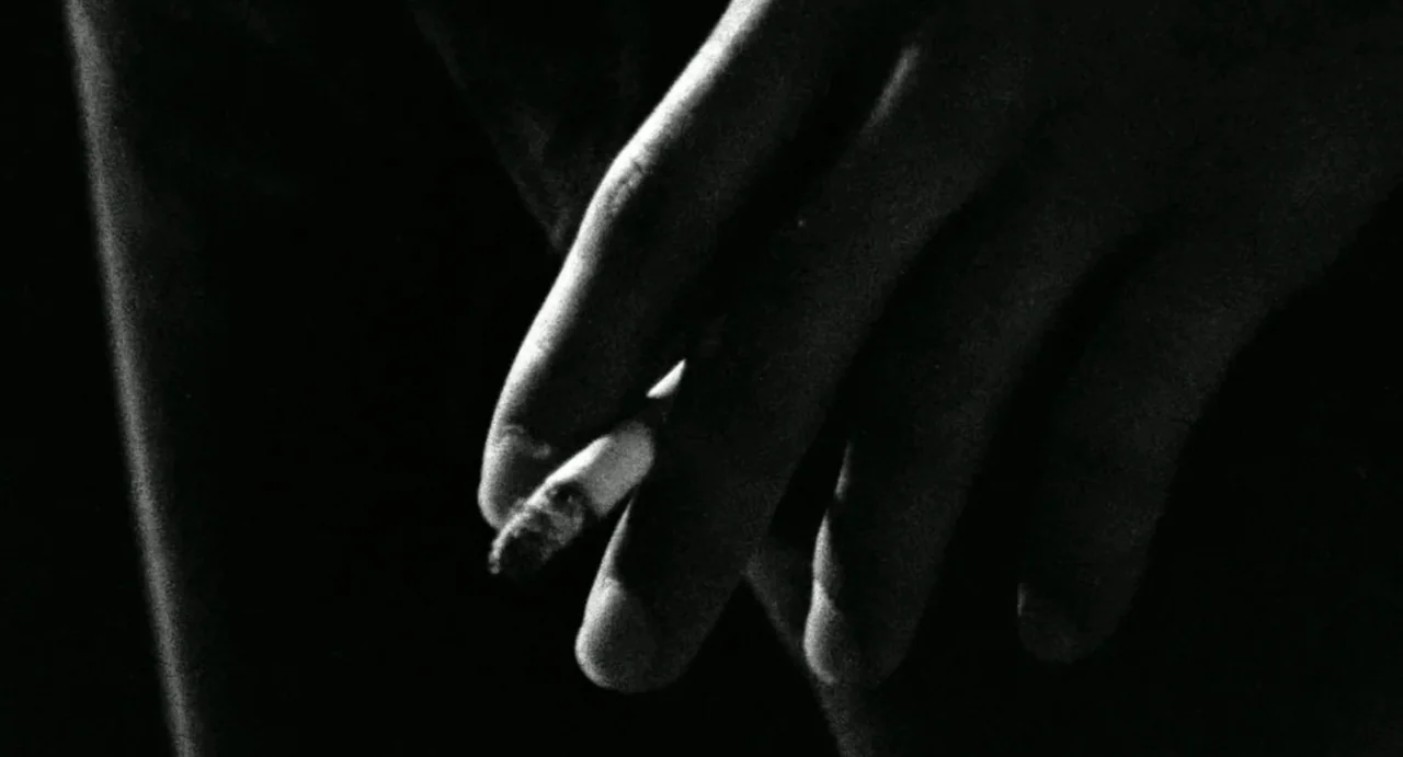 Burst City (1982) movie still: closeup — A close view of a hand holding a cigarette against a pitch black background. The cigarette is lit with…