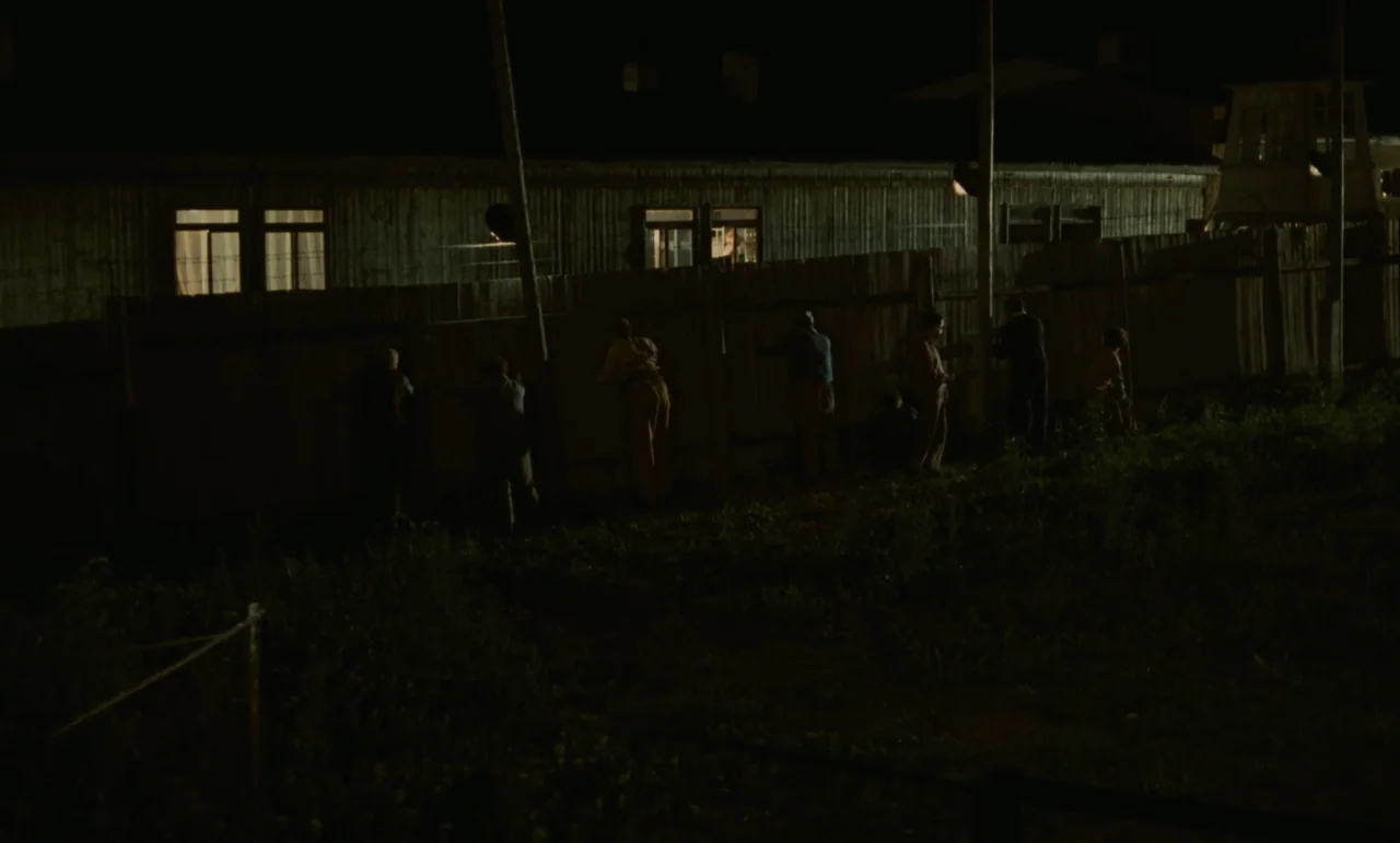 Larks On A String (1969) movie still: wide — A group of men stand lined up against a tall wooden wall at night. Harsh overhead lighting creates high…