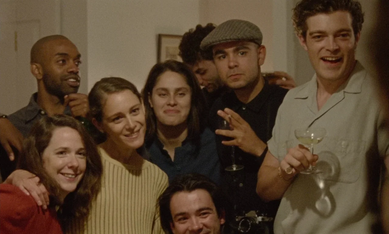 The Souvenir: Part II (2021) movie still: medium — An indoor gathering of seven young adults stands close together in a room. They smile toward the…