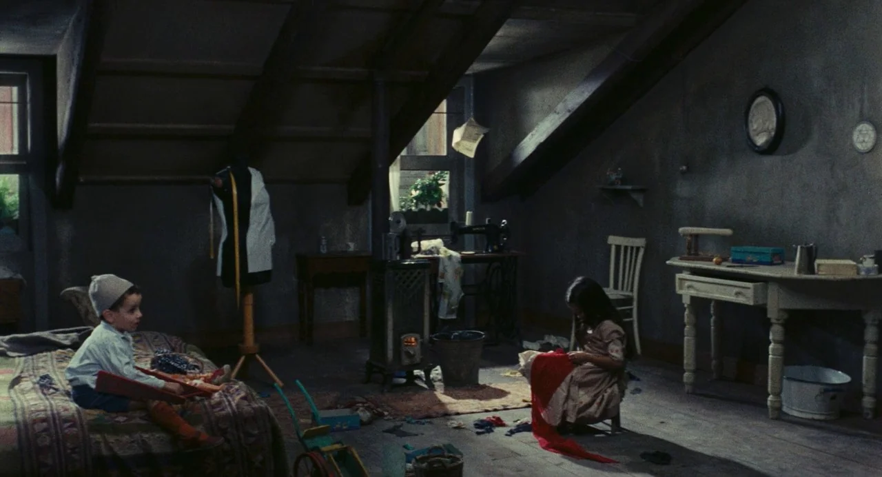 The Damned (1969) movie still: wide — A dimly lit attic features slanted wooden ceiling beams and cluttered period furniture. A girl sits on the…