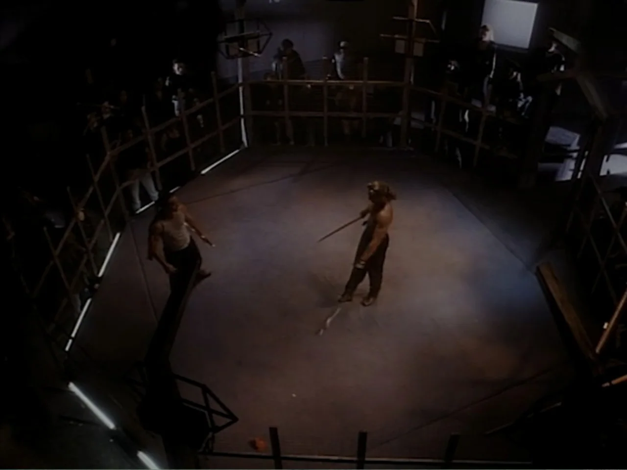 Firepower (1979) movie still: wide — An octagonal fighting ring surrounded by wooden barricades and observers in a dark room. Two shirtless men…