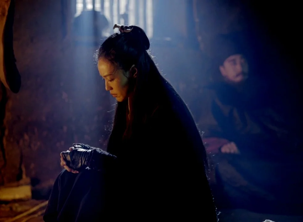 The Assassin (2015) movie still: medium — A dimly lit stone chamber with heavy shadows and a single barred window opening. A woman sits in the…