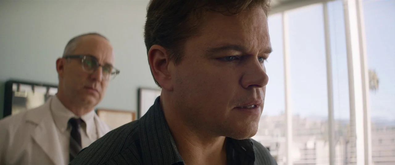 Ford V Ferrari (2019) movie still: medium — An office interior features a large window with white blinds and blurry city background. A man stands in…