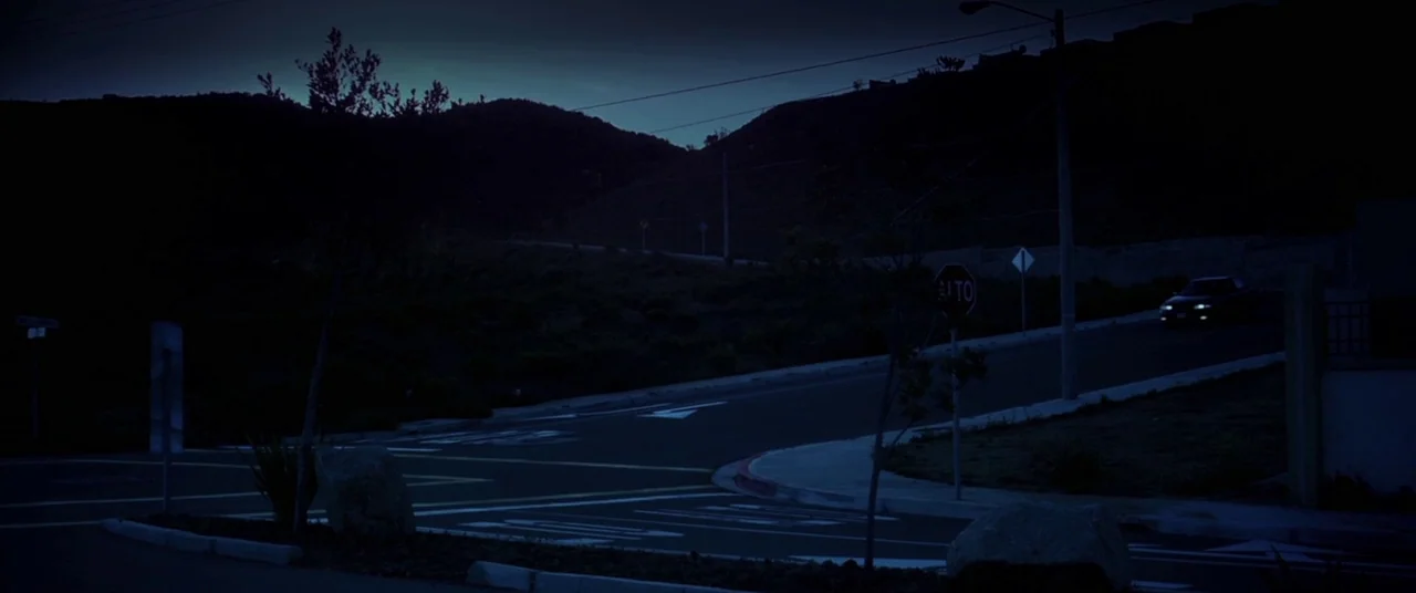 Here Comes The Devil (2012) movie still: wide — A dark, empty paved intersection sits under a clear night sky with rolling hills. A single car…