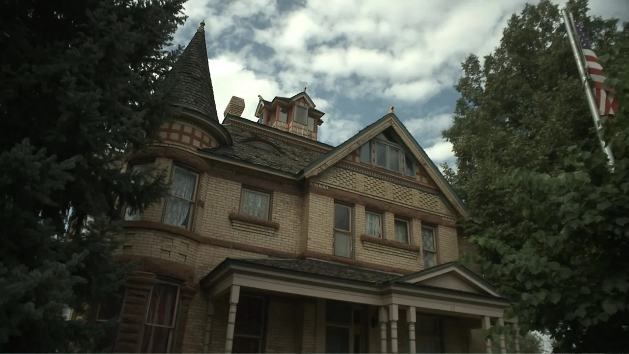 Boathouse Detectives (2010) movie still: establishing — A two-story yellow brick Victorian house with a conical roof tower stands outdoors. Large…