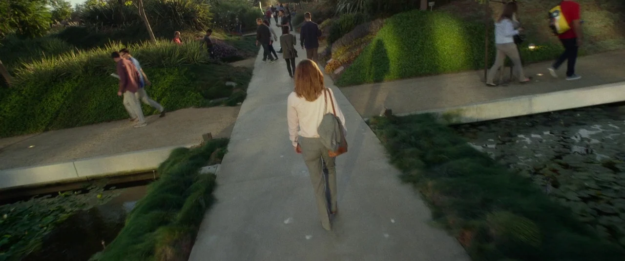 The Circle (2017) movie still: wide — A public park features a concrete walkway flanked by grassy mounds and a lily pond. Several pedestrians walk…
