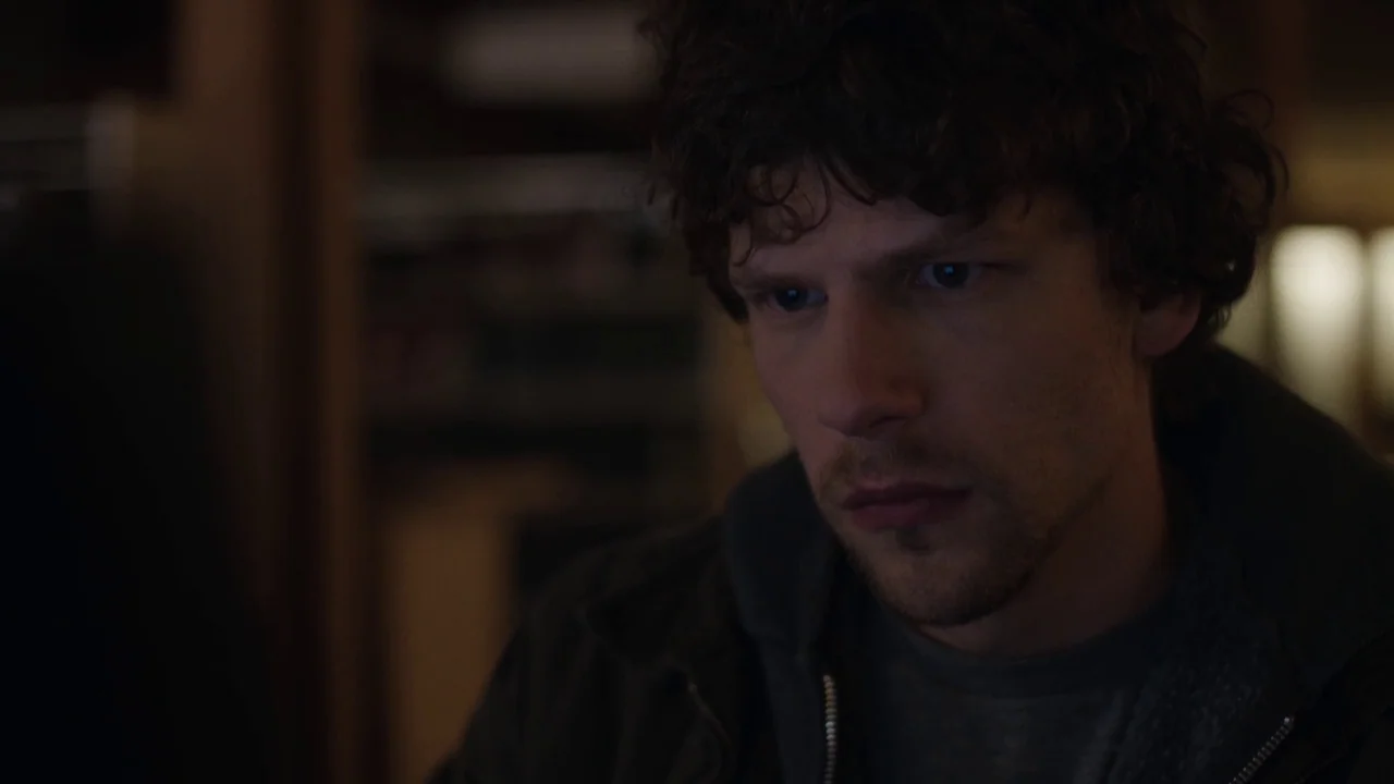 Night Moves (2013) movie still: closeup — A dimly lit room provides a dark background for the subject's face. A man with curly hair gazes off-camera…