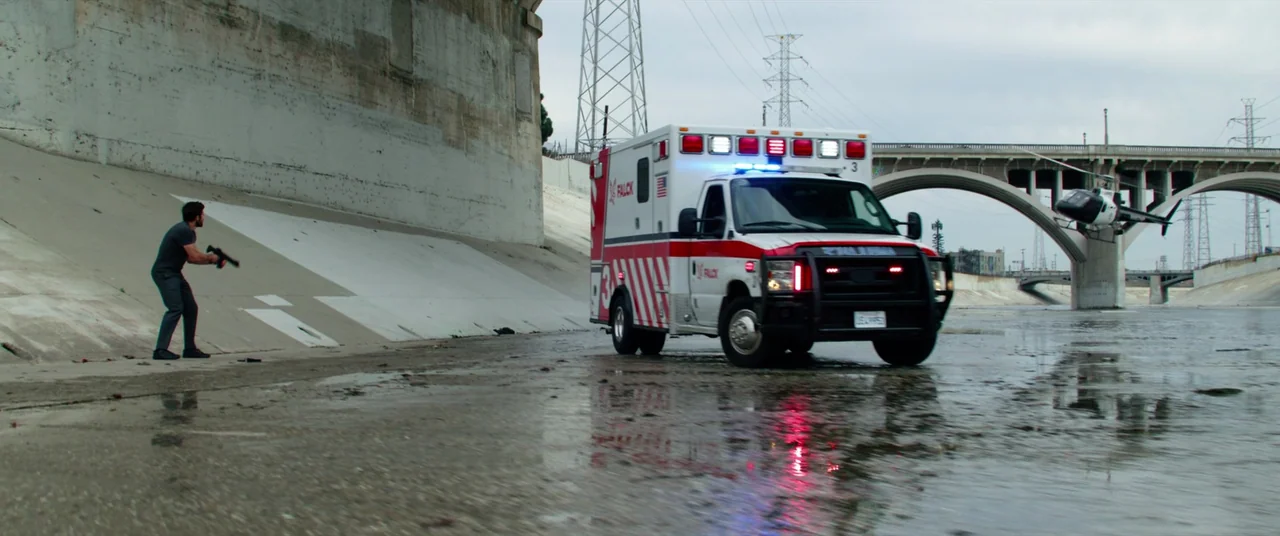 Ambulance (2022) movie still: wide — A wide shot shows a concrete storm drain with a man holding a firearm. An ambulance drives toward him while a…