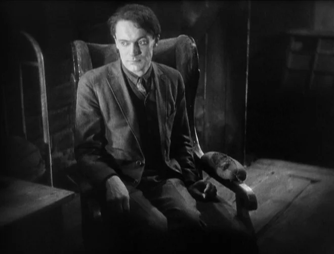 Pandora’s Box (1929) movie still: medium — A dark, sparsely furnished room with a wooden chair and wall textures visible. A man in a suit sits in a…