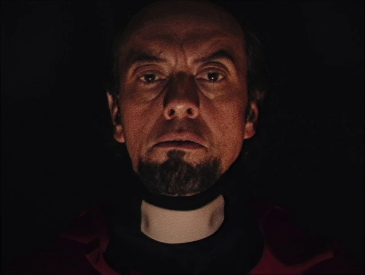 Late Night With The Devil (2023) movie still: closeup — A dark environment surrounds a man wearing a clerical collar. The man looks forward toward…
