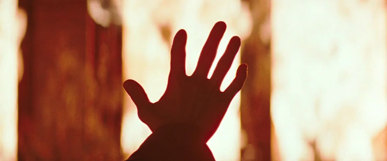 The Adjuster (1991) movie still: closeup — A close up view of a hand silhouetted against a wall of fire. Bright flames engulf the area behind the…