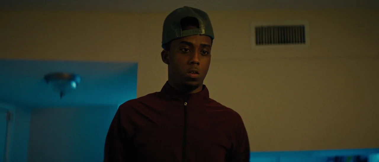 Moonlight (2016) movie still: medium — A residential interior space featuring a beige wall with a recessed air vent. A young man wearing a baseball…