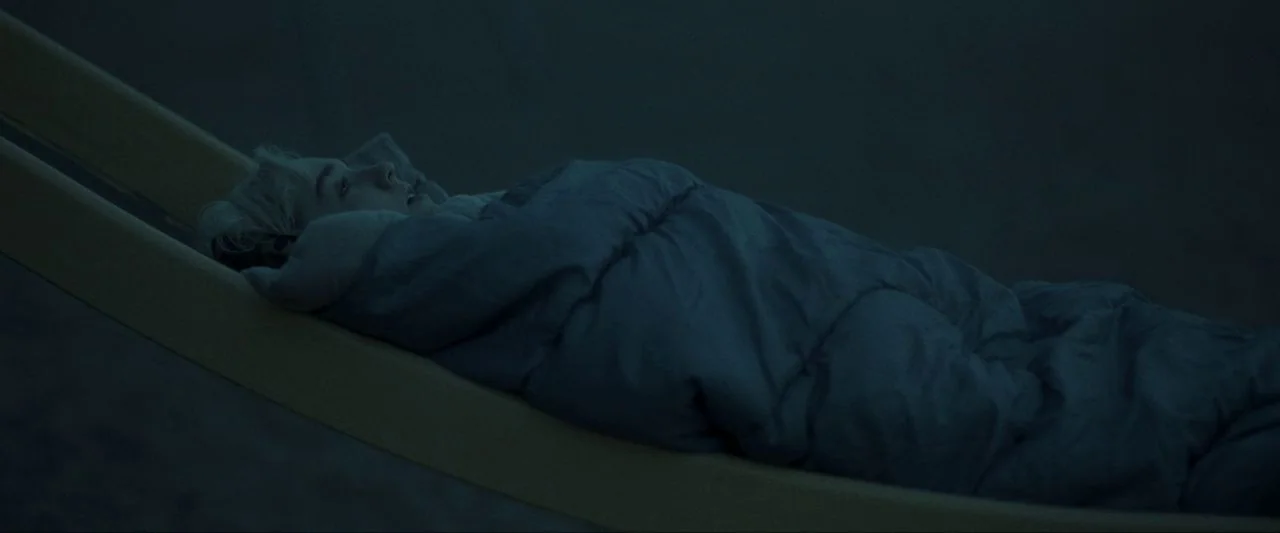 Come True (2020) movie still: medium — A person rests on a yellow metal slide during the dark of night. They are tightly wrapped in a sleeping bag…