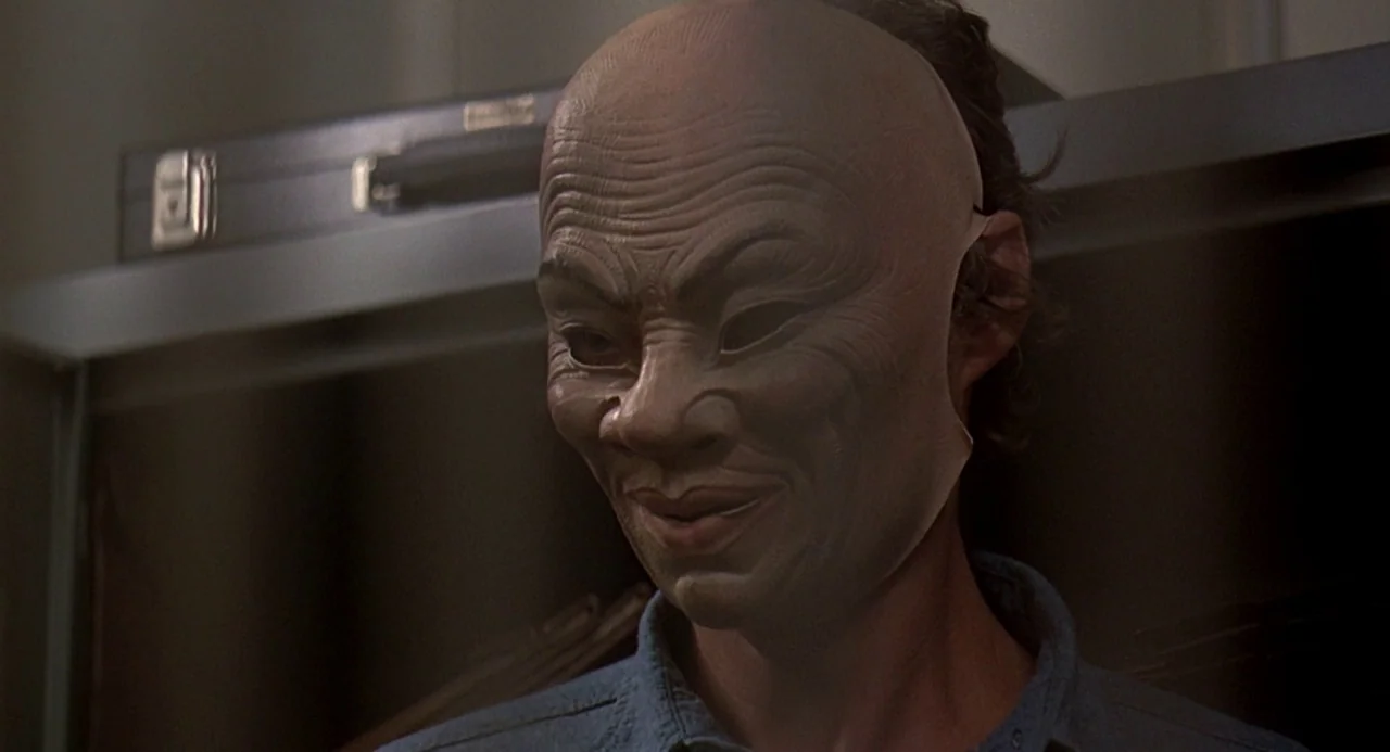 Charley Varrick (1973) movie still: closeup — A dark interior space features a person wearing a textured tan rubber mask. The individual is…