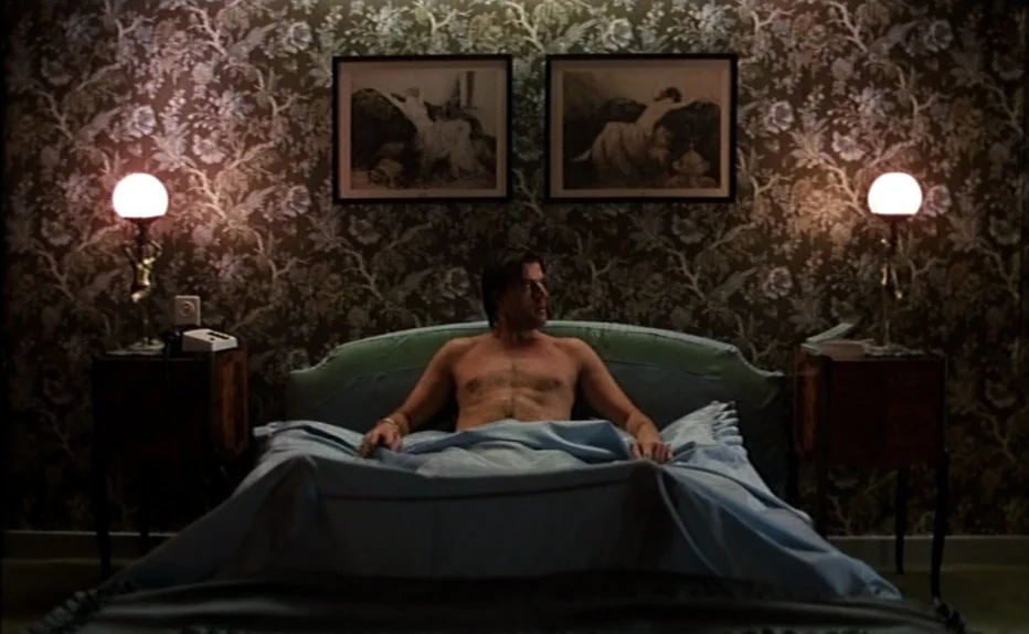The Fourth Man (1983) movie still: medium — An interior space features patterned vintage wallpaper and two framed prints above a bed. A shirtless…