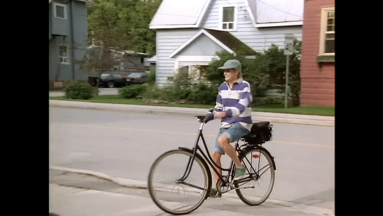 The Awakening (1995) movie still: fullbody — A quiet suburban street with houses and parked cars visible in the background. A teenager rides a black…