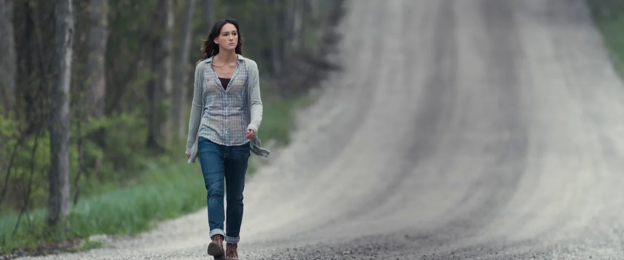 You’re Next (2013) movie still: fullbody — A woman walks down a wide, unpaved gravel road surrounded by a dense forest. The road curves into the…