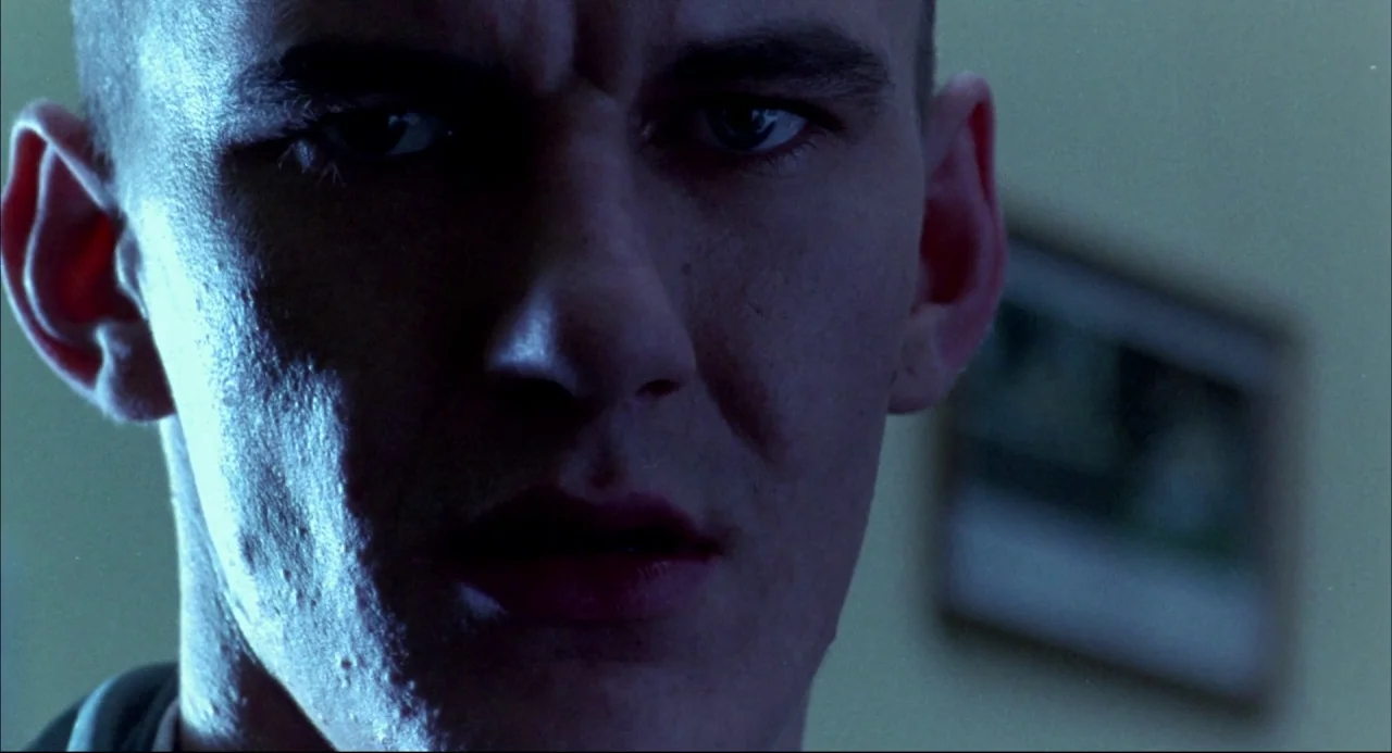 Romper Stomper (1992) movie still: closeup — A close portrait of a man in a dimly lit indoor space. The subject stares ahead with a tight and…