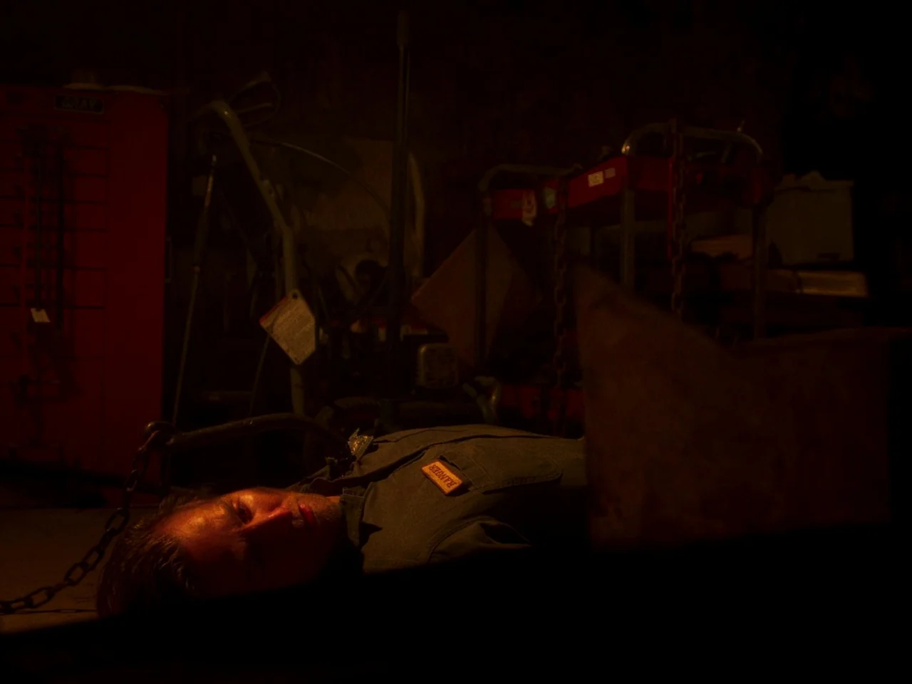 In A Violent Nature (2024) movie still: medium — A dark, cluttered industrial space with heavy tools and machinery visible in shadows. A man lies…