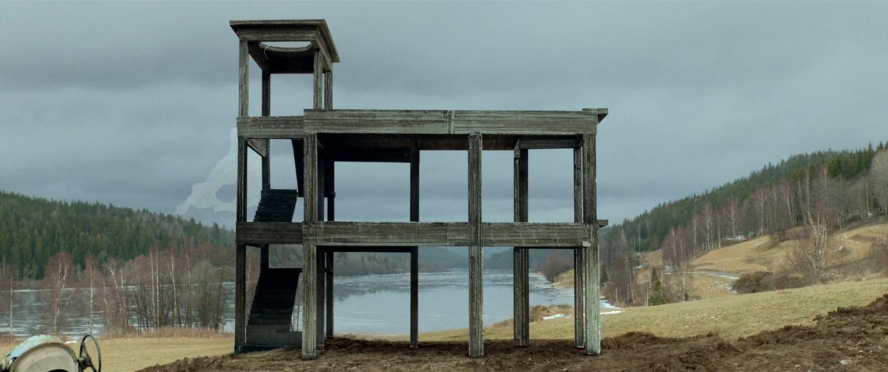 The House That Jack Built (2018) movie still: wide — A two-story concrete frame stands unfinished in a rural landscape near a river. Sparse…