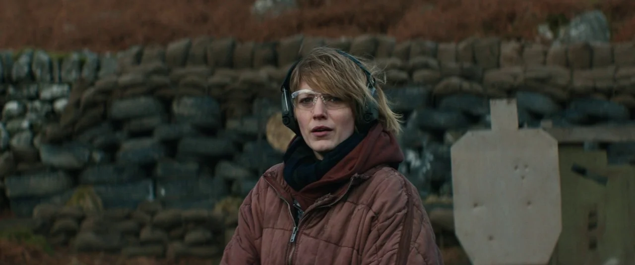 The Rhythm Section (2020) movie still: medium — A woman stands in front of a dirt embankment reinforced with stacked tires. She wears ear protection…