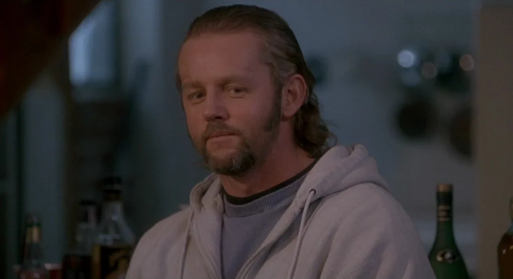 The Crossing Guard (1995) movie still: closeup — A dimly lit bar interior featuring blurry bottles on shelves in the background. A man with a goatee…