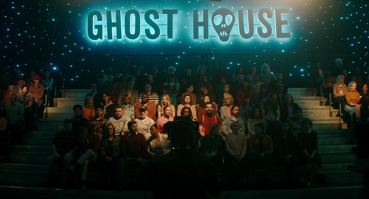 Beetlejuice Beetlejuice (2024) movie still: wide — An indoor auditorium with tiered seating is occupied by a large audience of people. A bright neon…