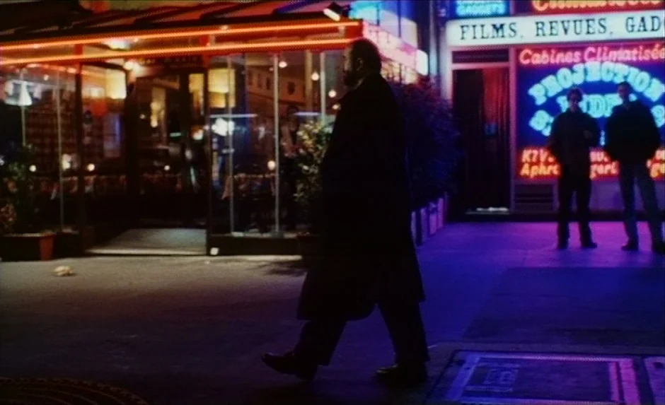 Simon, The Magician (1999) movie still: fullbody — A dark city street at night is illuminated by glowing neon storefront signs. A man in a dark coat…