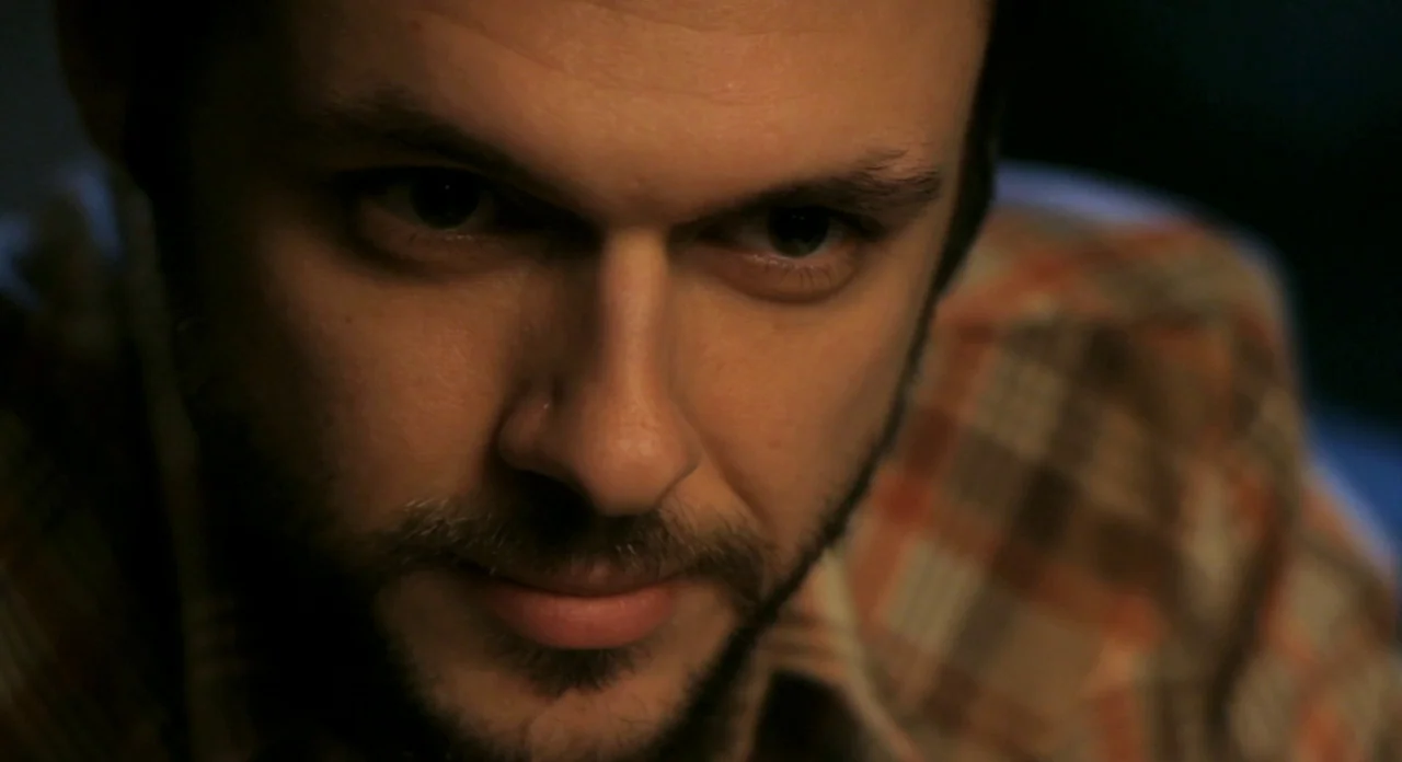 Sound Of My Voice (2011) movie still: closeup — A dark indoor space with minimal ambient light. A man stares forward from a close proximity to the…