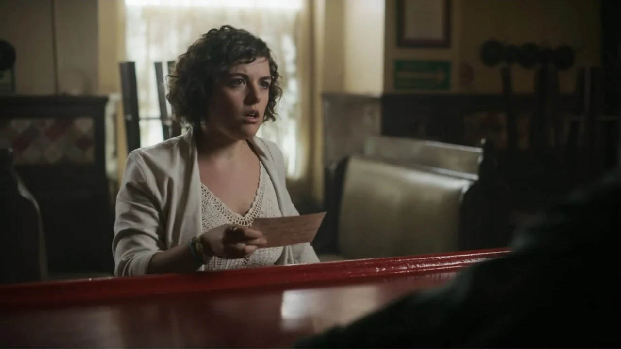 Lecciones para canallas (2022) movie still: medium — A dimly lit diner booth has a woman seated opposite an unseen conversational partner. The woman…